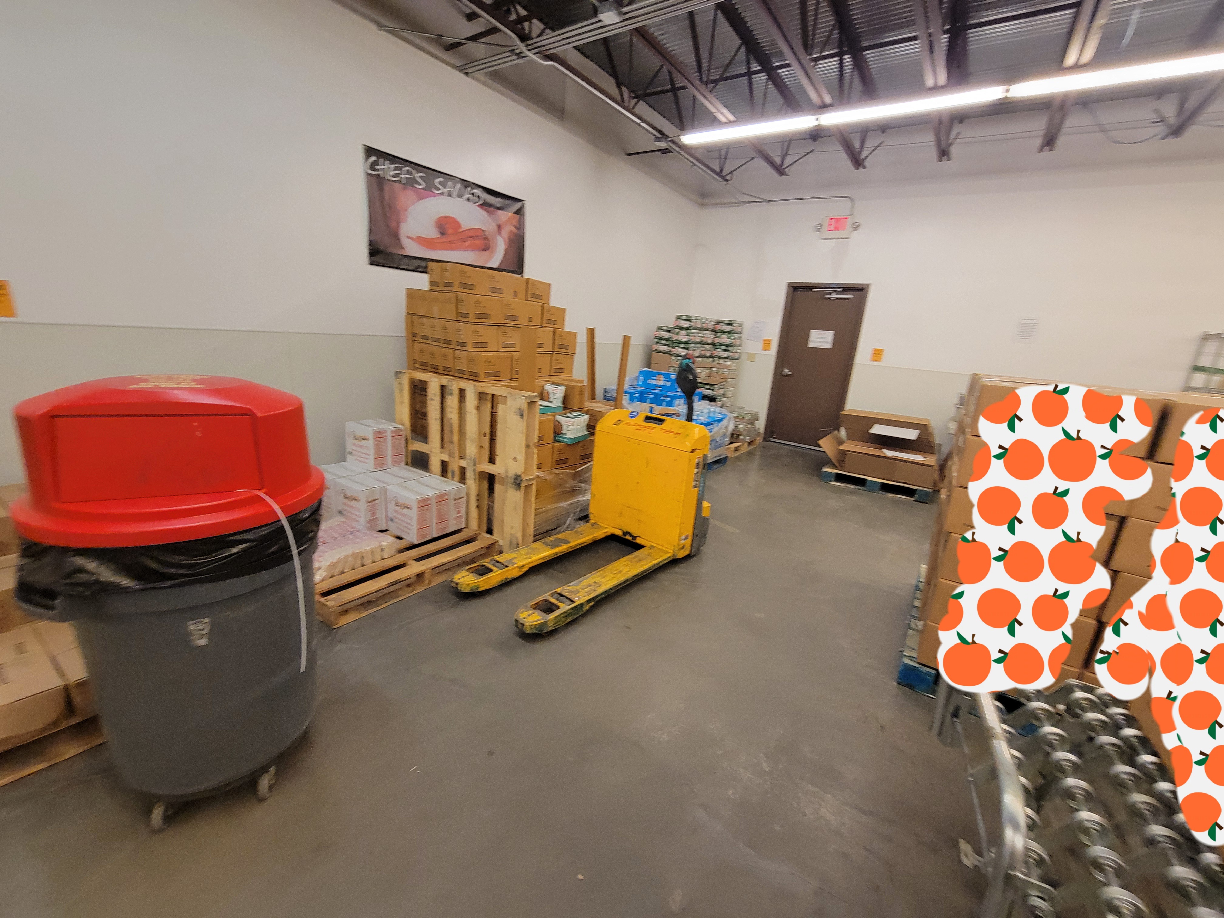 Pallets of food behind a pallet jacket and trash can. Next to tbose are a pallet of boxes and conveyor belt with a box on top.